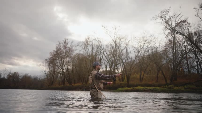 A man wearing a fishing vest and waders stands waist deep in a river casting his line against a backdrop of bare autumn trees and a cloudy sky