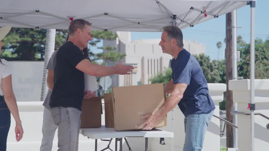 The video shows a group of diverse adults gathered under a white canopy on a sunny day likely participating in a community or volunteer eventa man in a blue shirt hands a large cardboard box to a man in a black shirt and the two briefly shake hands