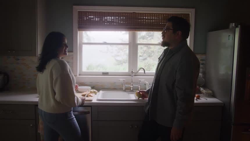 A woman and a man are standing facing each other by a kitchen sink under a large window with the woman momentarily reaching up to gently give food in the mans mouth on a kitchen setting