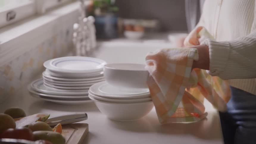 Woman standing up next to a counter with a sink drying dishes with a kitchen towel indoors during day time
