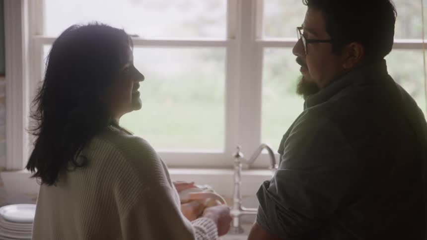 A man and a woman stand by a kitchen sink near a bright window drying a white plate and conversing happily