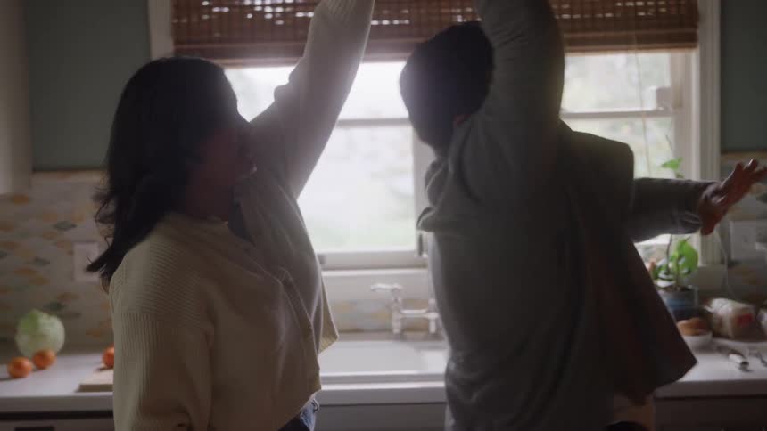 This video shows a couple dancing or swaying together in a warmly lit kitchen they are facing each other and in one frame they raise their arms above their heads while standing in front of a window with blinds