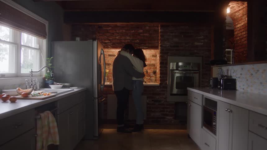 A couple is seen embracing and standing in a dark central corner of a kitchen featuring an exposed brick wall and gray cabinets while the figures are backlit and silhouetted by light coming from an upper window and a leftside window