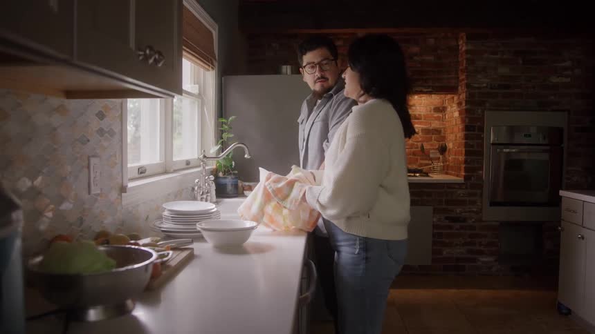Two people side by side drying dishes in kitchen with brick wall and natural light through window