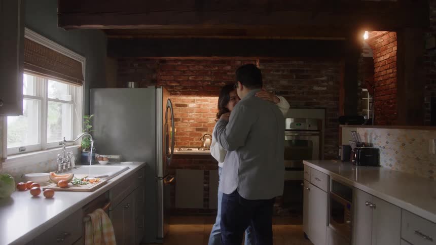 A man approaches a woman who is washing vegetables at a sink in a rustic kitchen with exposed brick and they cease the chore to embrace and then dance slowly together in the center of the room smiling and holding hands