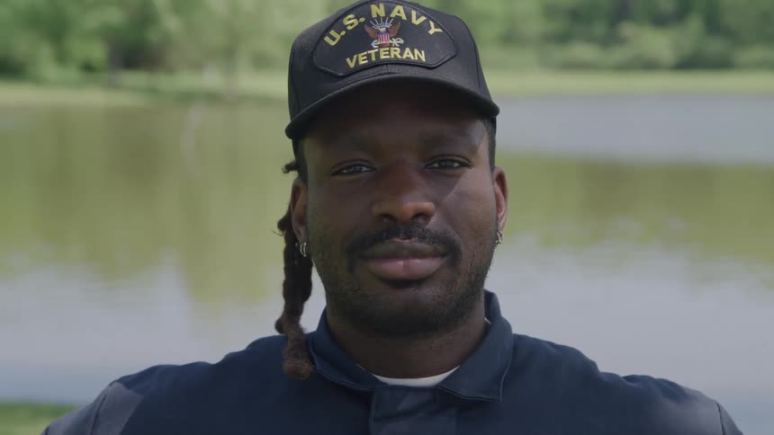 A man wearing a black us navy veteran baseball cap and a dark blue shirt looking directly at the camera with a calm expression with a blurred natural background of a body of water and green trees