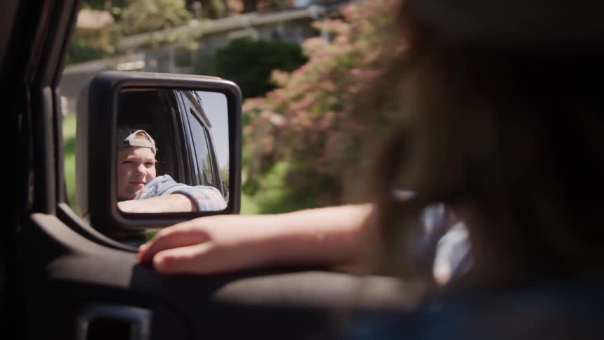 A teenager is looking in the side mirror of a vehicle as it moves down the road