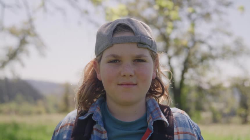 A young girl with long brown hair wearing a backwards baseball cap and backpack smiles at the camera in a sunny field outdoors