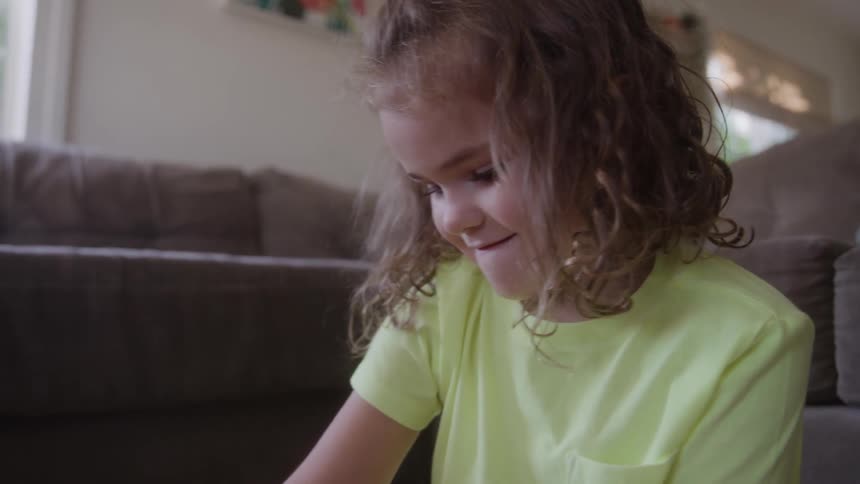 A little girl with curly brown hair wearing a light yellow shirt smiles happily while looking down in a sunlit living room