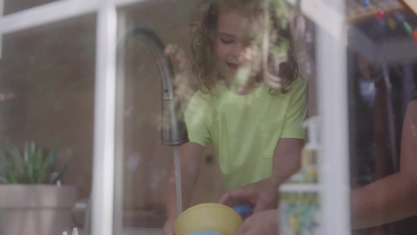 A mother and daughter wash utensils together at the kitchen sink