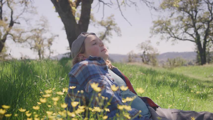 A young person possibly female with a peaceful expression looking forward while relaxing in a grassy field with yellow wildflowers trees and hills in the background