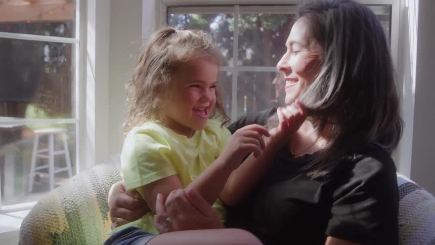A mother and her young daughter sit together on a sunlit sofa indoors sharing a playful and affectionate momentthe daughter wearing a yellow shirt laughs brightly while touching her mothers hair and the mother smiles down at her with joy