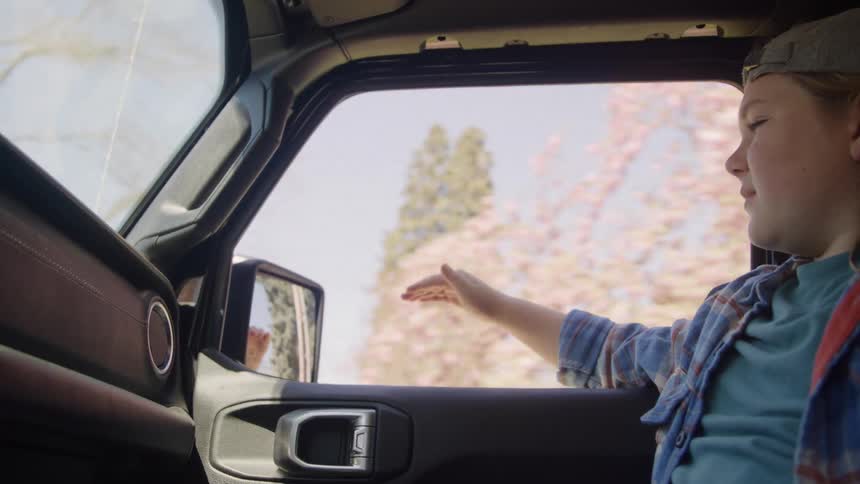 Child with backward cap extending arm out of moving vehicle among blossoms