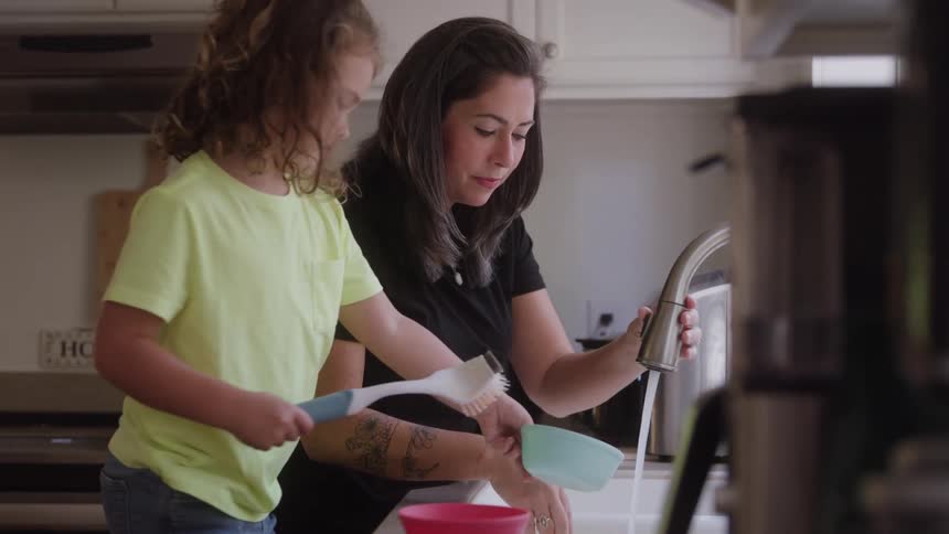 Mother and daughter washing plates inside the kitchen sink at dawn