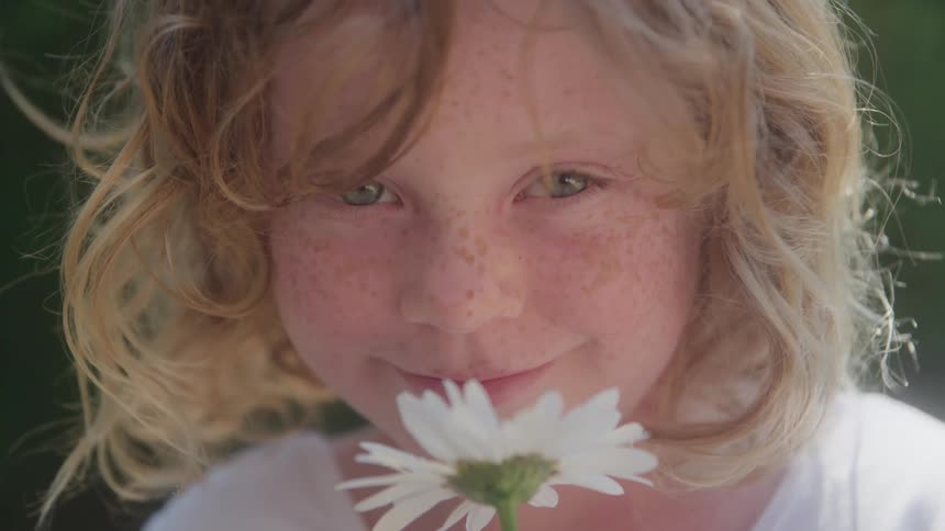 A closeup shot shows a young child with blonde windswept hair and freckles smiling slightly and looking directly at the camera while holding a white daisy up to the bottom of her nose
