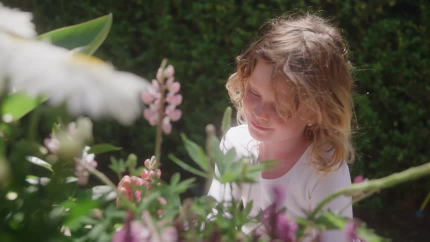 This video clip shows a young person with long sunlit wavy hair enjoying time in a gardenshe is wearing a white shirt and are surrounded by lush greenery and pink and purple flowering plants