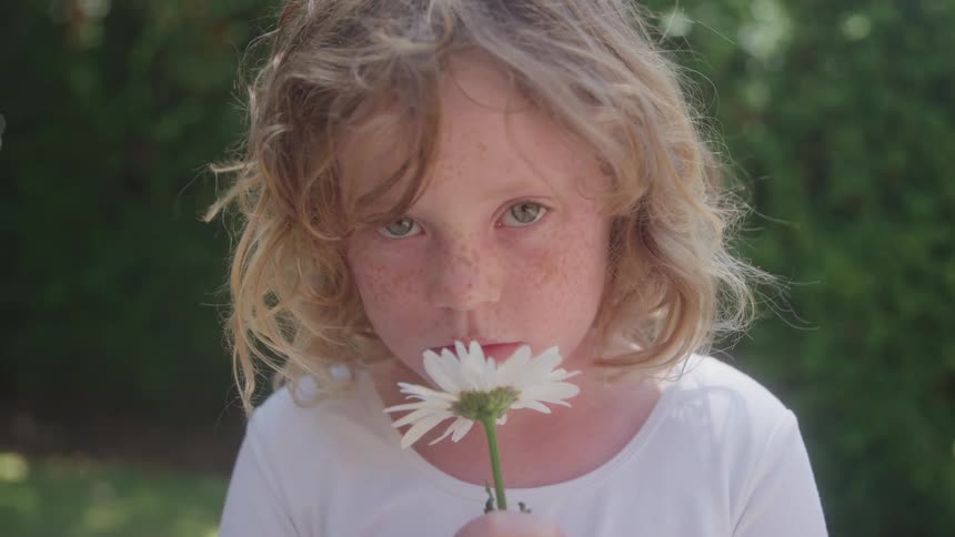 This is a closeup shot of a young child with freckles and wavy blonde hair smelling a white daisy flowerthe child is wearing a white shirt and is outdoors with green foliage visible in the softly focused background