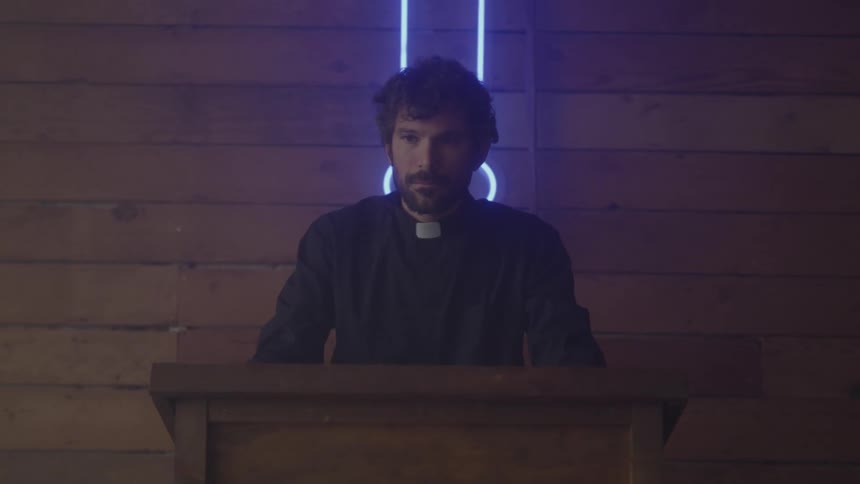A man in a dark priest or pastor shirt with a clerical collar stands behind a wooden podium illuminated by a neon cross on a wooden wall behind him