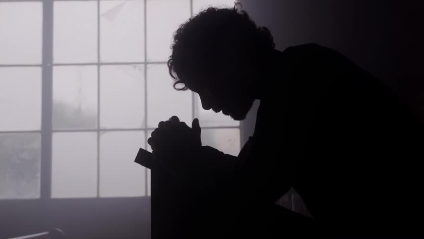 The video shows a silhouette of a person kneeling or sitting with their head bowed and hands clasped together possibly in prayer or deep contemplationthe strong backlighting comes from a large gridded window creating a somber highcontrast and dr