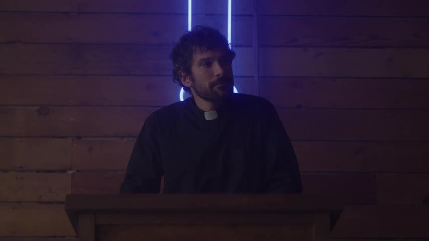A young pastor stands on the pulpit wearing a black shirt with a white clerical collar preaching to the congregation as a blue strip light glows behind him