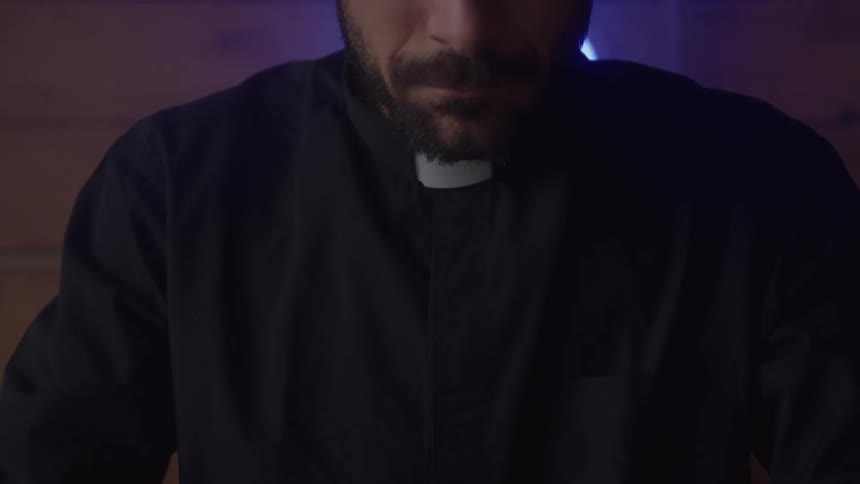 A close view of a man with a beard wearing a dark religious collar shirt looking down in a dimly lit room with wooden walls
