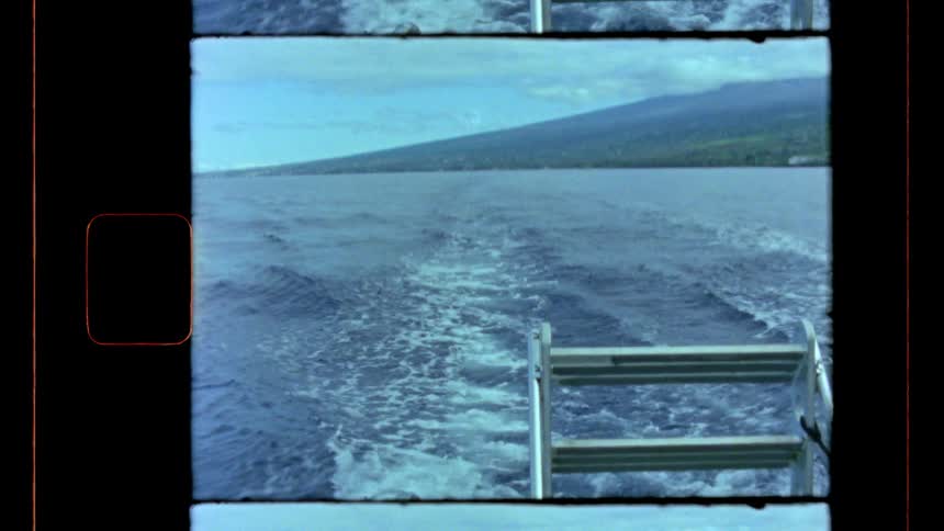 A low angle stern view from a boat shows the wake and a metal ladder as the vessel moves toward a large green mountain under a pale blue sky