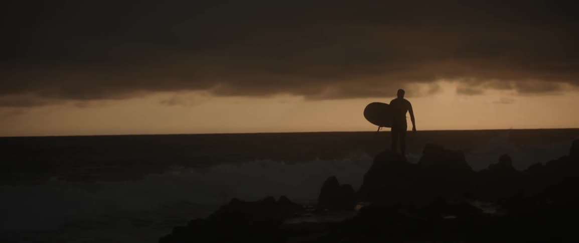 A man stands on rocks by the shore carrying a surfboard