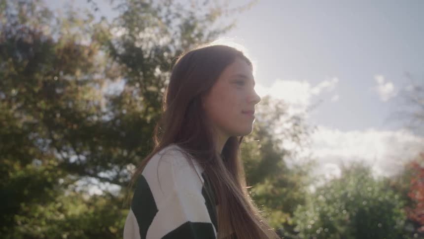 A teenage girl with long brown hair walks through the woods on a sunny day surrounded by tall trees