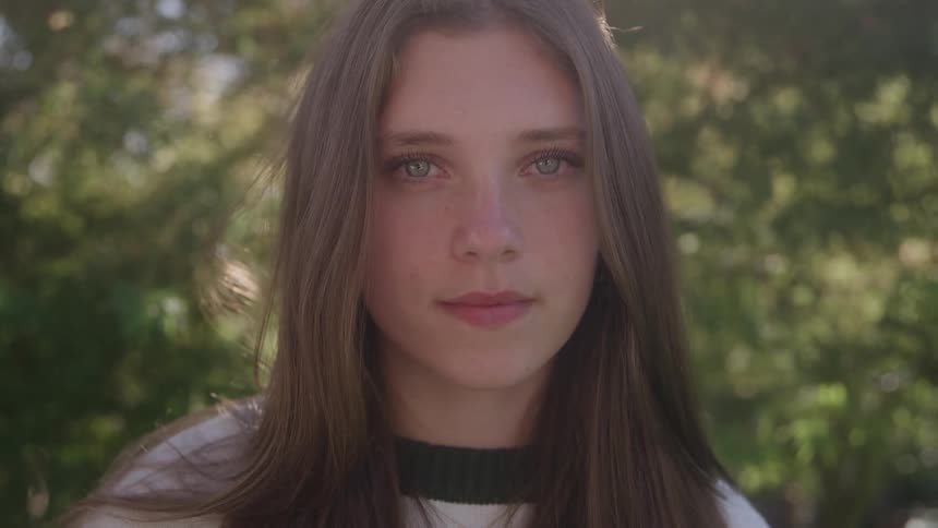 The video shows a closeup shot of a young woman with long brown hair and lightcolored eyes she is looking directly at the camera with a neutral expression and the background is blurred foliage suggesting an outdoor setting