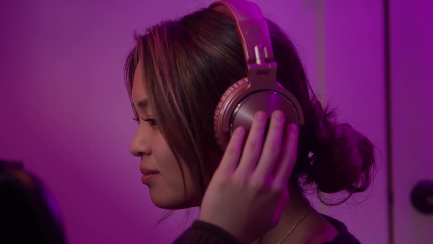 This is a closeup video of a woman wearing rose gold overear headphones adjusting them while listening to music the scene is bathed in a strong magenta or purple ambient light suggesting a latenight or studio setting