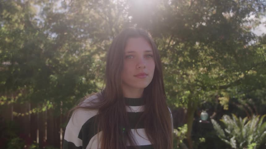 A young woman with long dark hair wearing a black and white striped shirt turning from a profile view to face the camera directly with a subtle smile standing outdoors in a bright sunny backyard