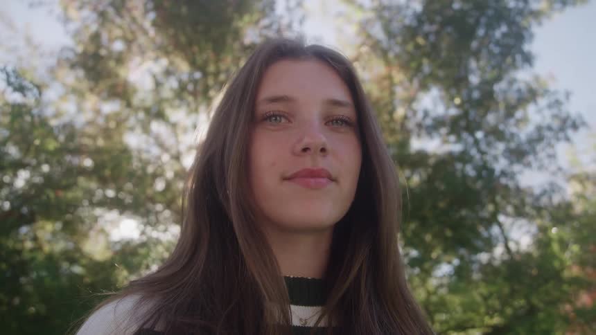 The video features a closeup of a young woman standing outdoors initially looking up and to the sideshe then turns to look directly at the camera and smiles with the sun briefly flaring over her face through the leaves of the trees behind her