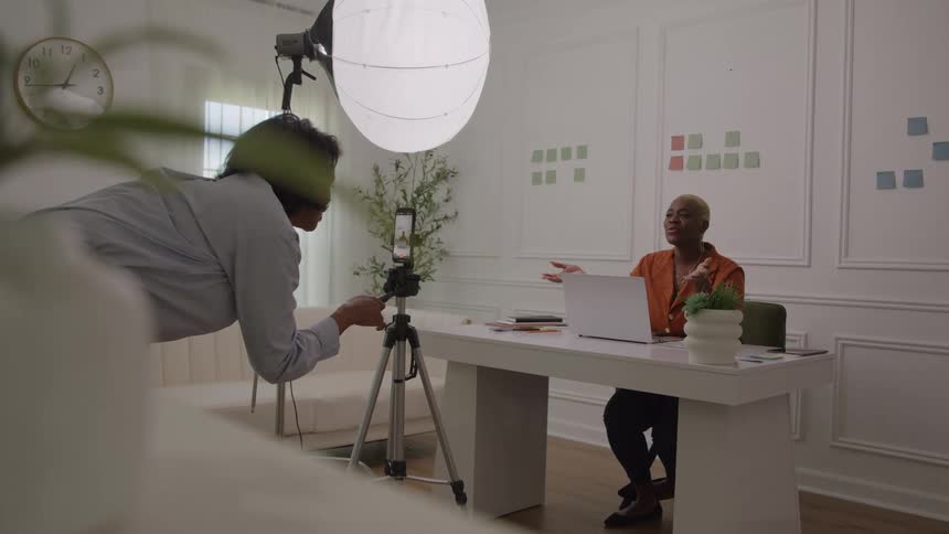 Young black woman talking and sitting on a chair in front of a laptop on a desk while another young black woman films her using a smart phone on a tripod indoors during day time