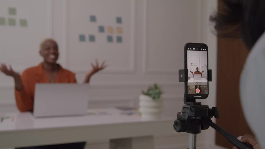 A smartphone mounted on a small tripod in the foreground is recording a smiling person with short blonde hair wearing an orange shirt who is sitting at a desk with a laptop and gesturing with both hands in the background