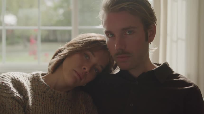 A young couple sits together on a couch the woman resting her head on the mans shoulder both look sad as sunlight beams softly through the window behind them