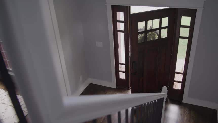 Viewed from the top of a staircase a woman opens her front door to receive a cardboard package then steps back inside a home entryway