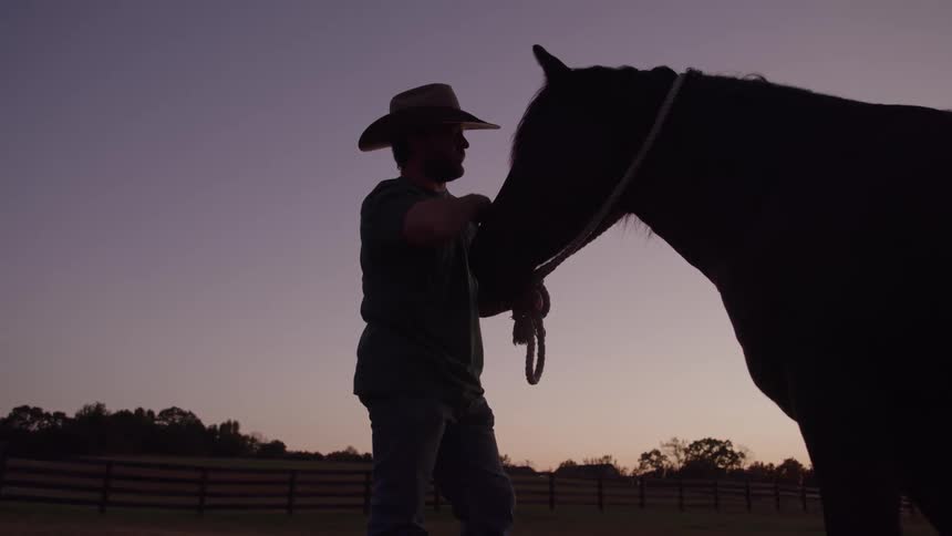 Silhouette of person in cowboy hat petting horse at sunset in fenced pasture
