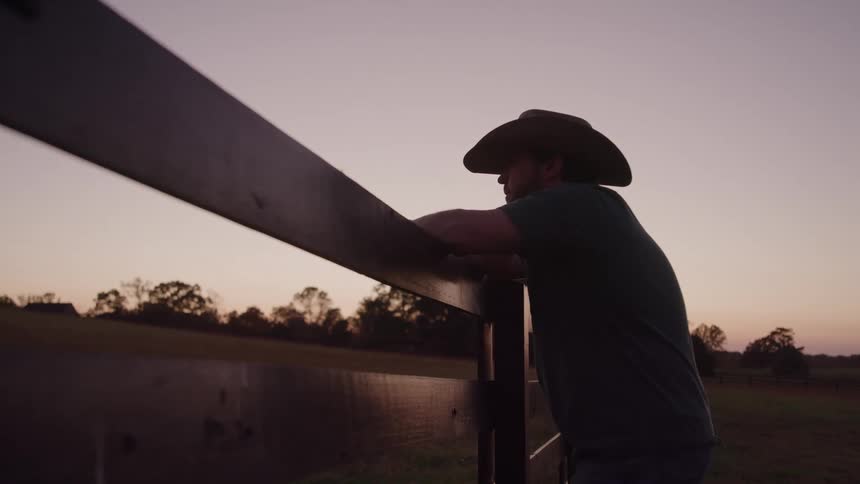 This is a shot of a man wearing a cowboy hat leaning against a dark wooden fence at sunset or sunrisethe man is silhouetted against the colorful sky suggesting a moment of quiet contemplation in a rural setting