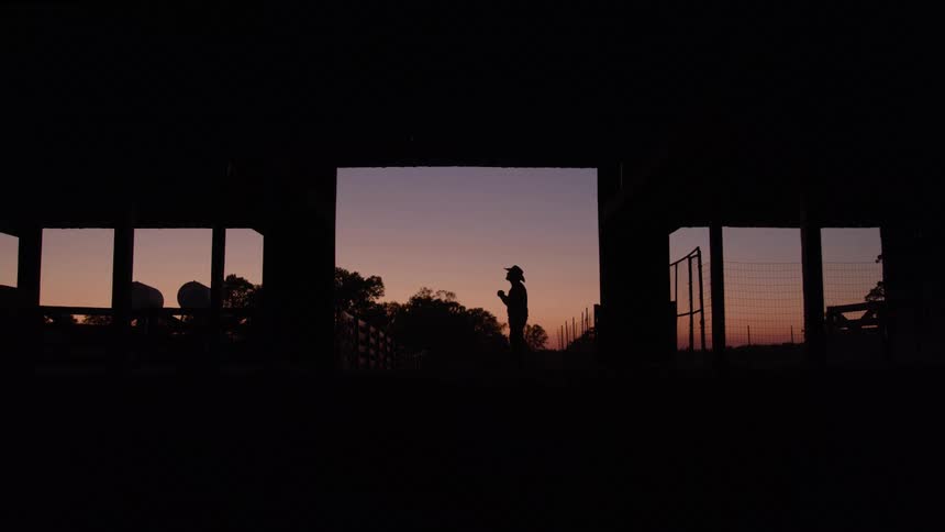 This is an interior shot looking out through a large framed doorway and windows at sunset or sunrisethe interior is in deep shadow strongly silhouetting a person wearing a hat who is standing outside in the center frame against the fading light