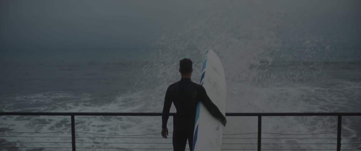 A man holds a surfboard as  waves crash against the shore