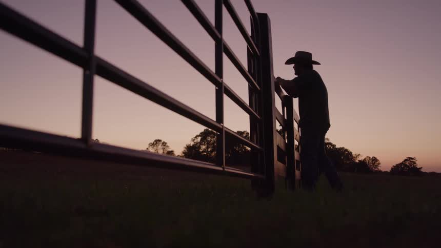 Silhouette of person in cowboy hat leaning on metal gate at sunset in rural setting
