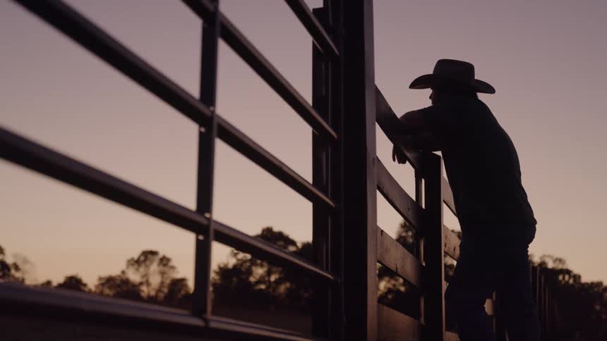 Silhouette of a man in a cowboy hat walking to a wooden fence and leaning on it for a while outdoors during dusk or dawn