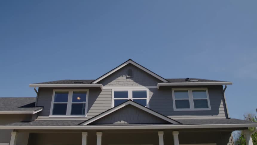 A slow tiltdown shot descends from a clear blue sky to reveal the front facade of a twostory grey suburban house with white columns