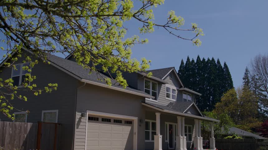Low angle shot looking up at bright green tree branches against a blue sky slowly tilting down to reveal a two story suburban home with a garage