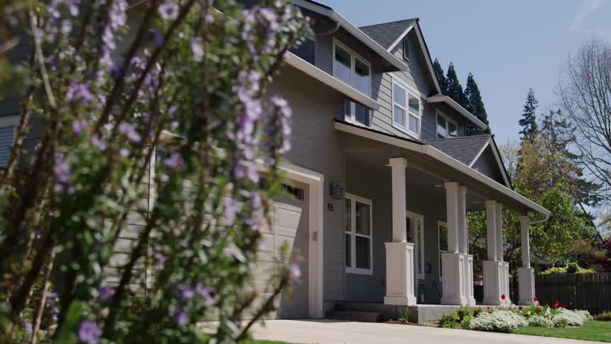 A dolly shot of a modern gray suburban home with a white columned porch and large garage framed by purple flowering shrubs on a sunny day