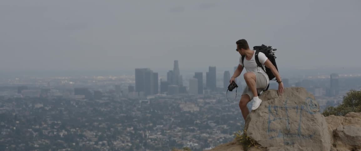 A man holding a camera on a mountain above a large city on a cloudy day