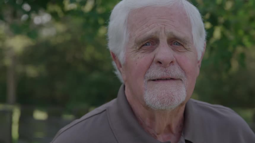 Older person in brown shirt in front of foliage lit by filtered sunlight