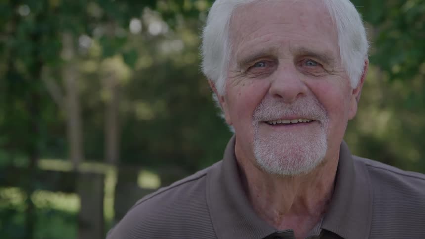 A closeup shot features an elderly man with bright blue eyes white hair and a goatee standing outdoors the man turns to face the camera slowly smiling warmly in a seemingly comfortable and relaxed setting
