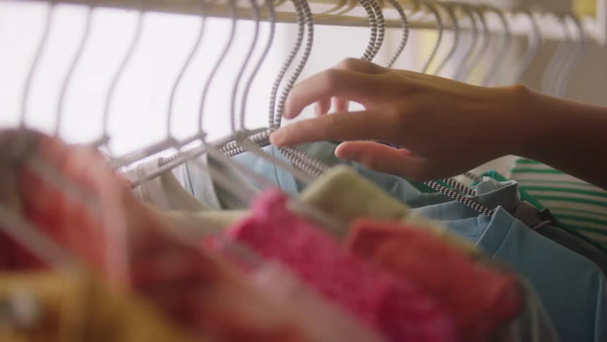 A close view of a womans hands sorting through a rack of colorful clothing hanging on wire hangers in a bright closet space