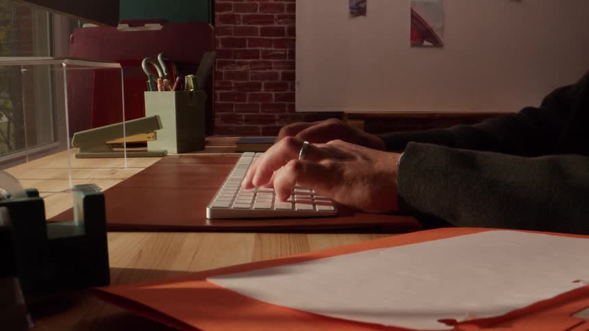 This video features a closeup shot of someones hands typing quickly on a white modern computer keyboardthe person is working at a desk that has office supplies a brown leather pad and stacks of papers and folders in the foreground and background